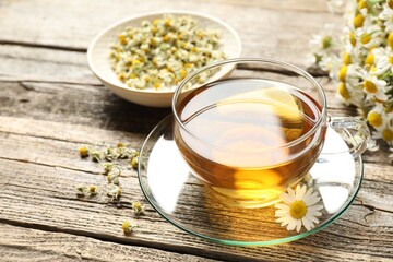 Aromatic tea in glass cup and chamomile flowers on wooden table, closeup