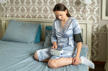 Woman checking reading on blood pressure monitor while sitting on bed, monitoring health at home, wellness and prevention concept