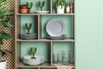 Shelving unit with different dishware, glasses and floral decor near green wall indoors