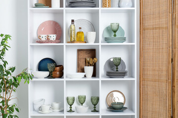 Shelving unit with different dishware, glasses and other kitchen utensils near white wall indoors