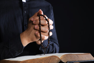 Priest with rosary and Holy Bible praying on black background, closeup. Space for text