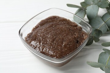 Natural body scrub in glass bowl and eucalyptus branches on white wooden table, closeup