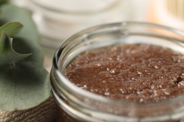 Natural body scrub in glass jar, eucalyptus leaves and towel on blurred background, closeup