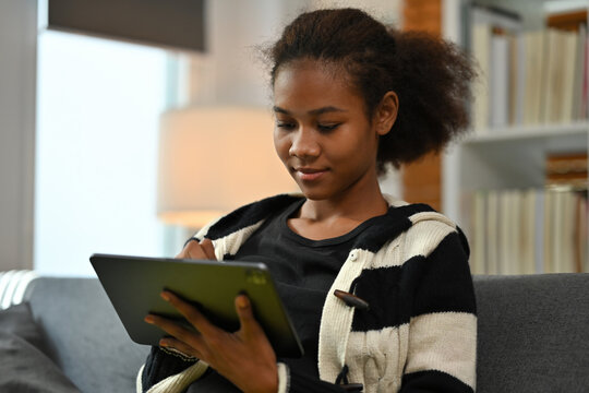Focused young African woman sitting on a sofa, using a digital tablet with a stylus in a cozy home - Powered by Adobe