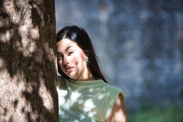 A beautiful Latin teenager with long dark hair rests her hands and face on a tree trunk in the...
