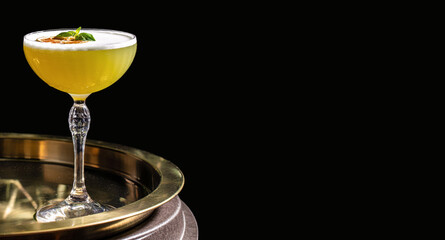 Yellow cocktail with a frothy foam top, garnished with dried citrus and basil leaf, served in an...