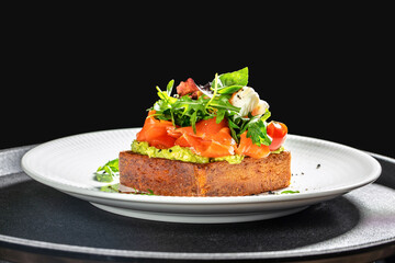 Gourmet open sandwich with smoked salmon, avocado spread, cherry tomatoes, fresh greens, and soft cheese served on a thick slice of rustic bread on a white plate