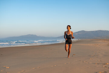 Smiling athletic woman running barefoot on the beach during sunrise, wearing a sporty black outfit, with ocean waves and distant mountains in the background on a peaceful morning.