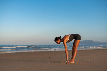 Young athletic woman stretching hamstrings on a beach at sunrise, performing a forward bend with...