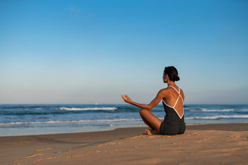 Fit woman meditating in lotus position on sandy beach at sunrise, wearing a black fitness outfit,...