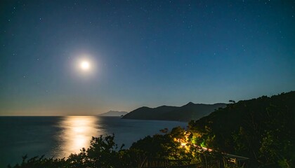 Fototapeta premium A tranquil coastal scene at night, showcasing a bright moon reflecting on the still water, with distant mountains and a winding road illuminated by lights below.
