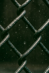 Detailed closeup of a metal chain link fence with water droplets, showing texture, strength, and a gritty atmosphere in a dark background.