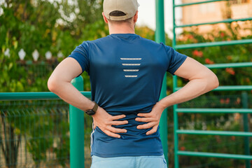 Man in blue athletic shirt stretches and rubs lower back pain during outdoor workout at fitness park in evening light