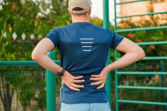 Man in blue athletic shirt stretches and rubs lower back pain during outdoor workout at fitness park in evening light