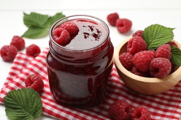 Tasty jam, fresh ripe raspberries and leaves on white wooden table, closeup