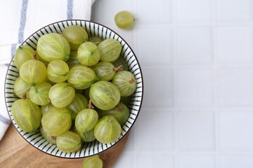 Fresh green gooseberries in bowl on white tiled table, top view. Space for text