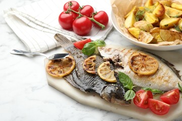 Tasty roasted flounder fish served with potatoes, tomatoes, basil and lemon slices on white marble table, closeup. Homemade seafood dish
