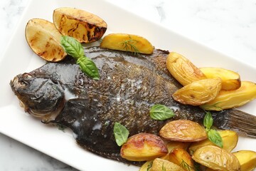 Tasty roasted flounder fish served with potatoes, basil and lemon slices on white marble table, top view. Homemade seafood dish
