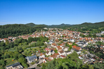 A panoramic view of the town of Dahn in the Pfälzerwald forest, showing the red-roofed buildings nestled in a green valley with a prominent rock formation behind it, under a clear summer sky.