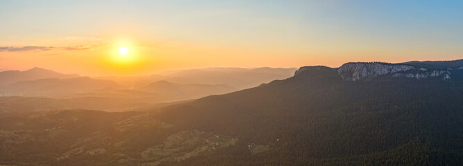 The sun slips behind the horizon, casting warm light across a vast mountainous skyline and forested slopes