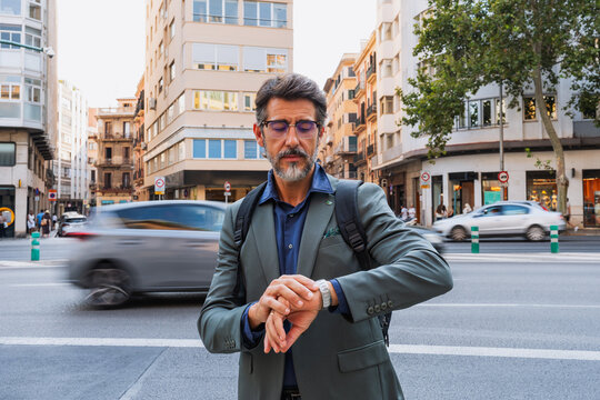 Stylish middle-aged man in suit looking at watch while standing on a busy street. Concept of business, time management, urban lifestyle