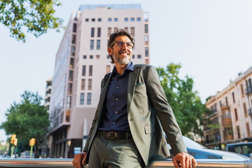 Confident middle-aged man in suit smiling in front of modern buildings. Concept of business, optimism, urban lifestyle