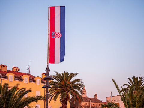 Croatian national flag on pole in coastal town with palm trees and historic Mediterranean architecture at sunset. - Powered by Adobe