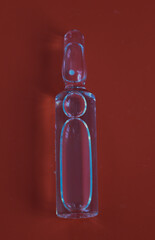 ​A vertical shot of a transparent medical ampoule lying on a red background. Light passing...