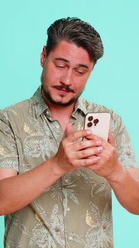Middle-aged man scrolls smartphone casually, then suddenly hits forehead with palm realizing he forgot something important. Young adult guy isolated on blue background showing memory lapse and urgency
