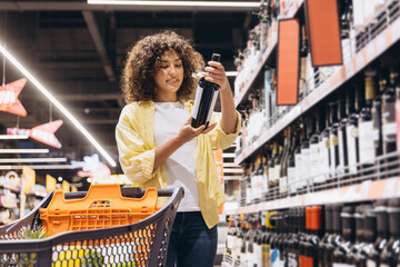 Woman choosing wine in supermarket drinks aisle pushing shopping cart