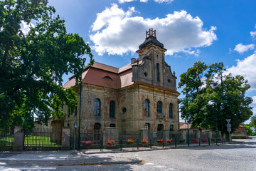 Obraz premium Baroque church in Goszcz surrounded by trees and fencing, located by a cobblestone street. Historic sacred architecture showcasing the monumental character of the heritage temple in Lower Silesia.