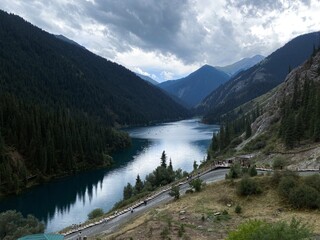 Blue waters of Kolsay lake among pine forest. Wooden pedestrian stairs below, majestic mountains above