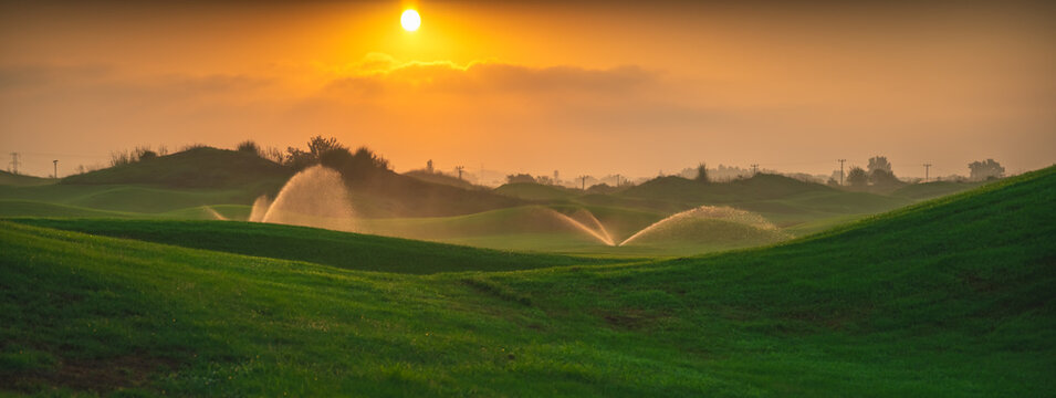 Automatic water sprinklers irrigating a lush green golf course at sunset, creating a serene landscape with golden light illuminating water droplets and rolling hills - Powered by Adobe