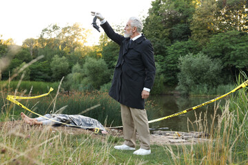 Forensic expertise. Detective with evidence working at crime scene with dead man's body near lake...