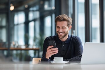 A smiling businessman in black shirt with a laptop using his smartphone in coworking space, with coffee cup and modern interior in the background. A man smiles while looking at his phone at a cafe.