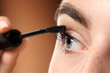 Young woman applying mascara onto her eyelashes on brown background, closeup