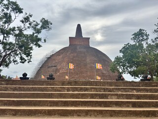 Majestic ancient brick stupa with colorful Buddhist flags fluttering against a cloudy sky, viewed...