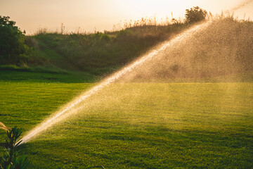 Water sprinkler spraying refreshing mist over lush green grass on a golf course during sunset,...