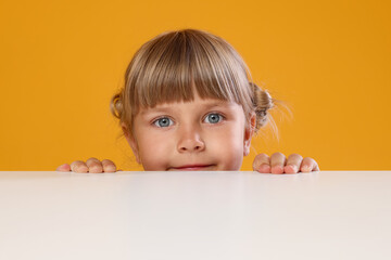 Curious little girl peeking over table against orange background