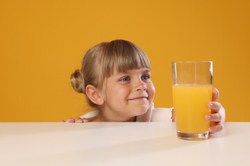Cute little girl reaching for glass of juice at white table against orange background