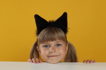 Curious little girl in cat ears peeking over white table against orange background