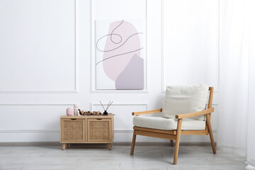Comfortable beige armchair and wooden cabinet with decor near white wall indoors