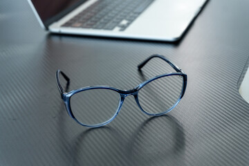 Eyeglasses with a blue frame sit on a sleek desk next to a laptop, highlighting a modern and organized workspace perfect for productivity
