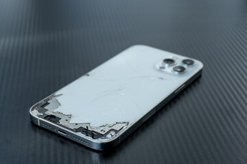 A smartphone with a cracked back and exposed internal parts is laid on a dark textured surface. The...