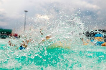 A wave of turquoise water crashes forward, sending droplets flying. In the background, people enjoy...
