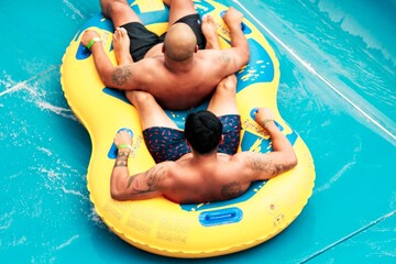 Two men wearing swimming trunks are on a yellow and blue inflatable raft sliding down a vibrant...