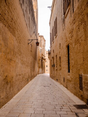 Historic cobblestone alley in Mdina, Malta, flanked by aged stone buildings and lanterns, leading...