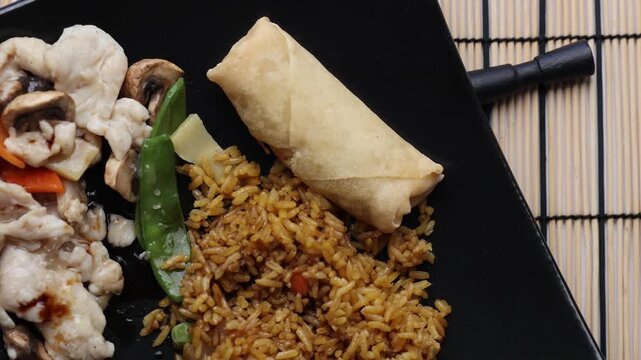 Extreme close-up of Moo Goo Gai Pan with fried rice and egg roll on a black plate, as metal chopsticks pick up rice, followed by pieces of meat and vegetables.