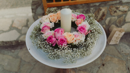 A stunning centerpiece features a white candle encircled by delicate pink and white roses, harmonizing with lush greenery. Perfect for a tranquil outdoor gathering, this arrangement radiates elegance.