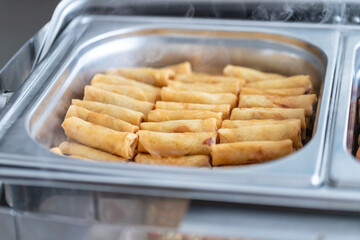 Savory spring rolls, perfectly fried to a crisp golden brown, steam gently in a stainless steel tray at a lively urban food festival. Aromas fill the air, inviting visitors to indulge.